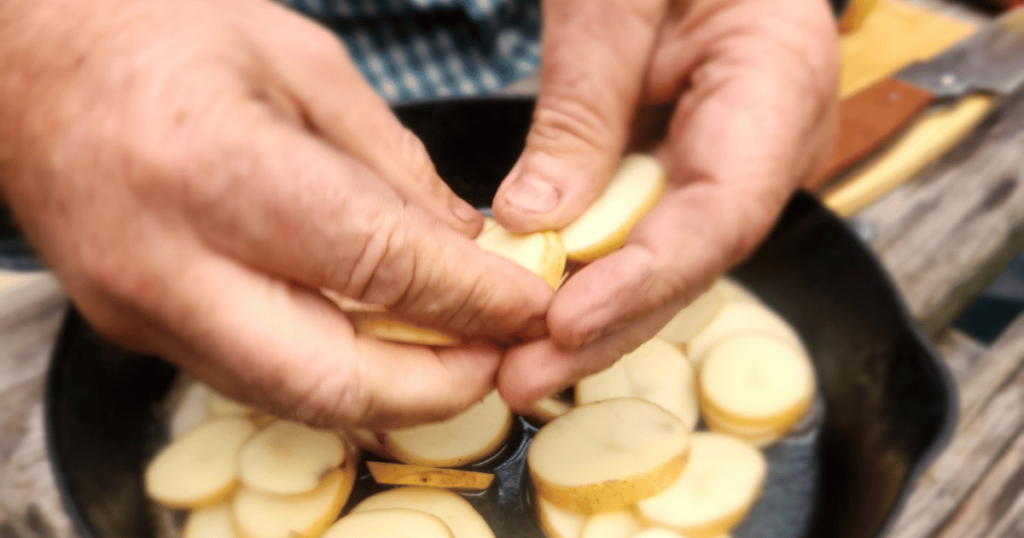 QUICK & EASY CAMPING SIDE DISH IDEAS FOR OUTDOOR ADVENTURES man placing sliced potatoes in a skillet at camp