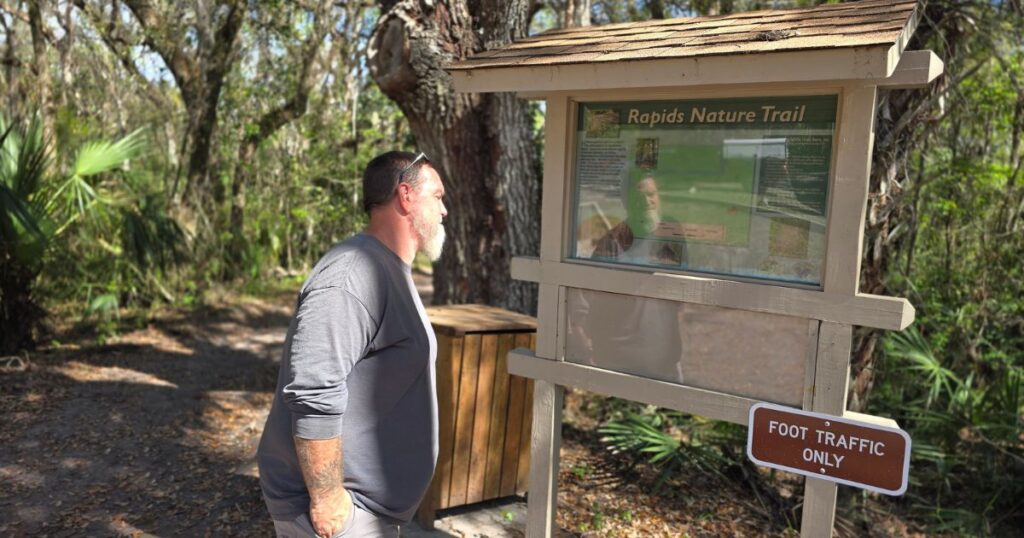 THIS IS AN IMAGE OF A MAN READING THE TRAIL HEAD SIGN FOR THE RIVER RAPIDS TRAIL