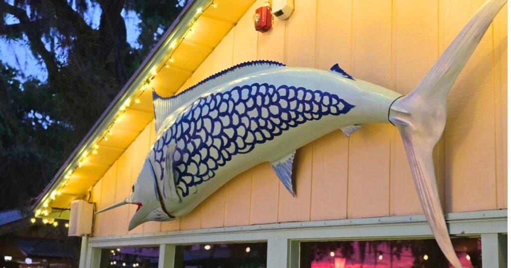 Large decorative fish mounted on the exterior wall of a waterfront restaurant in St. Augustine, lit by string lights at dusk.