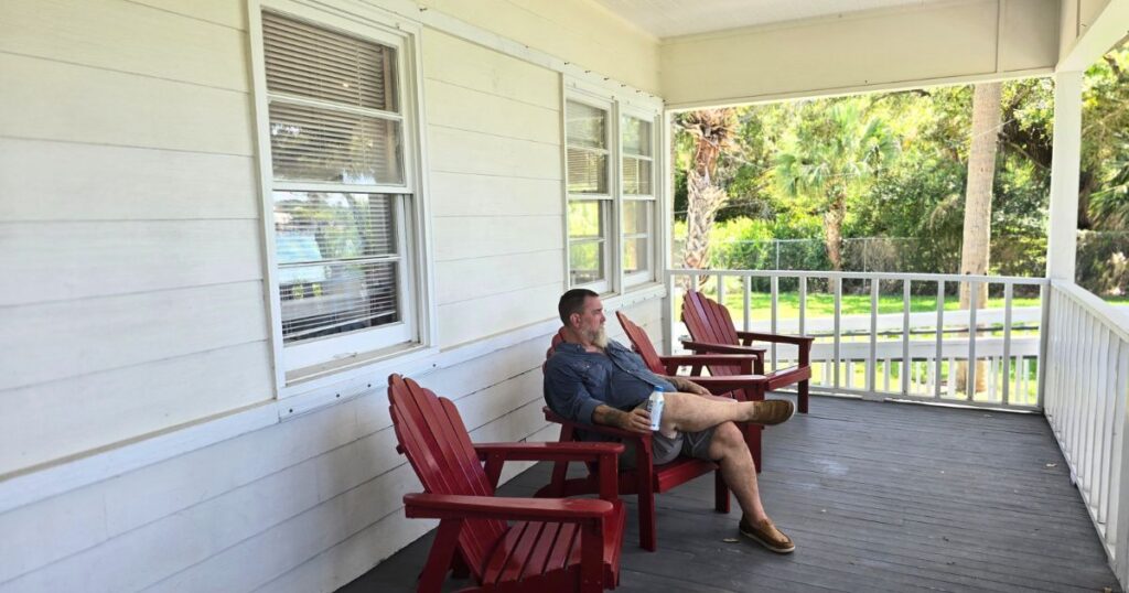 Relaxed man sitting on Red Adirondack chair on porch overlooking lush green garden.