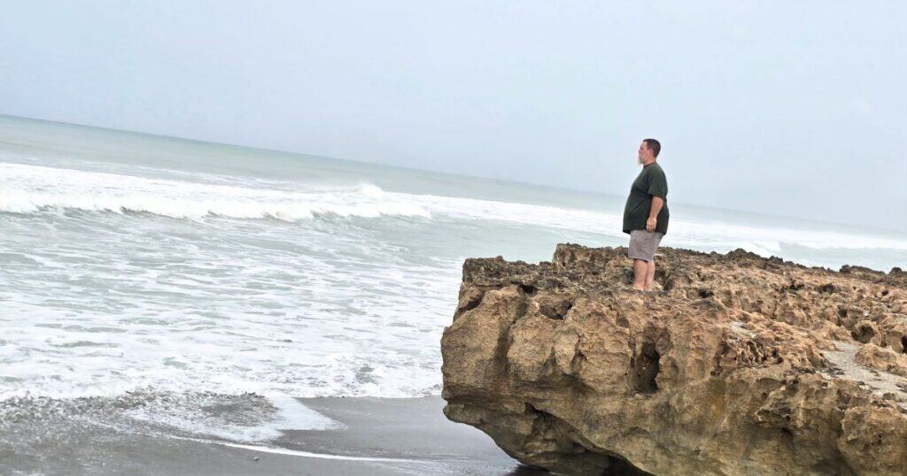 Seaside landscape with man standing on rocky shoreline, ocean waves crashing, overcast sky, Hawaii coast, contemplative moment.