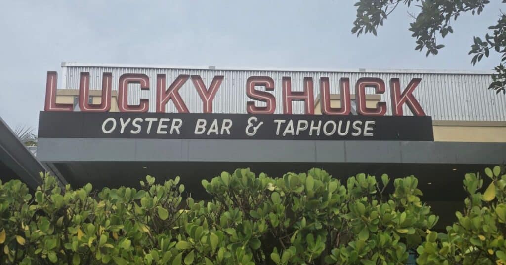 Lucky Shuck Oyster Bar and Taphouse sign, tropical foliage in foreground, inviting seafood restaurant and bar atmosphere.