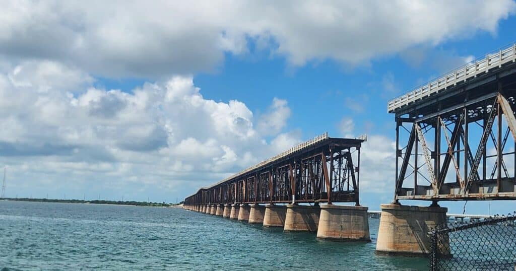 Old iron railroad bridge over water under blue sky with clouds, scenic coastal view.