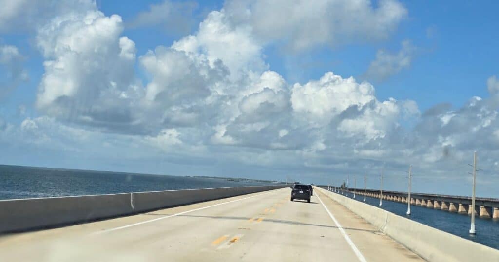 Seaside highway view with cars driving over a bridge, blue sky, and clouds, featuring The Wandering Hulasquatch theme.