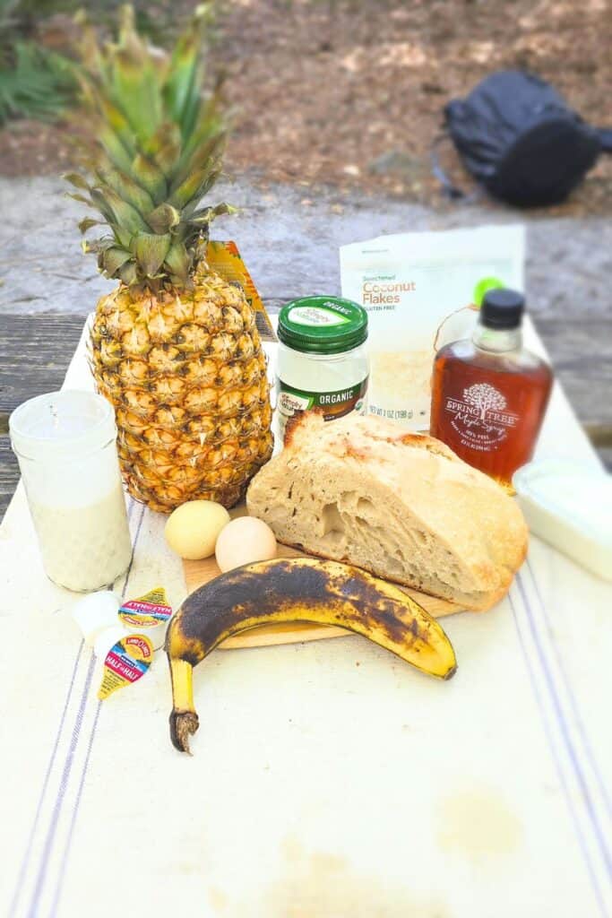 this is an image of ingredients on a picnic table for pina colada French toast. The image includes bread, banana, coconut milk, maple syrup, shredded coconut, and a whole pineapple.